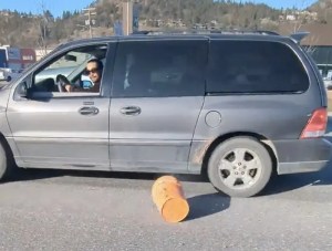 A man with sunglasses driving a grey van after throwing stuff out the window, and an orange bucket can be seen on the road.