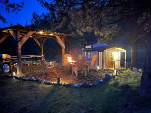 Wooden Adirondack chairs are places around a flaming fireplace in the woods at dusk.