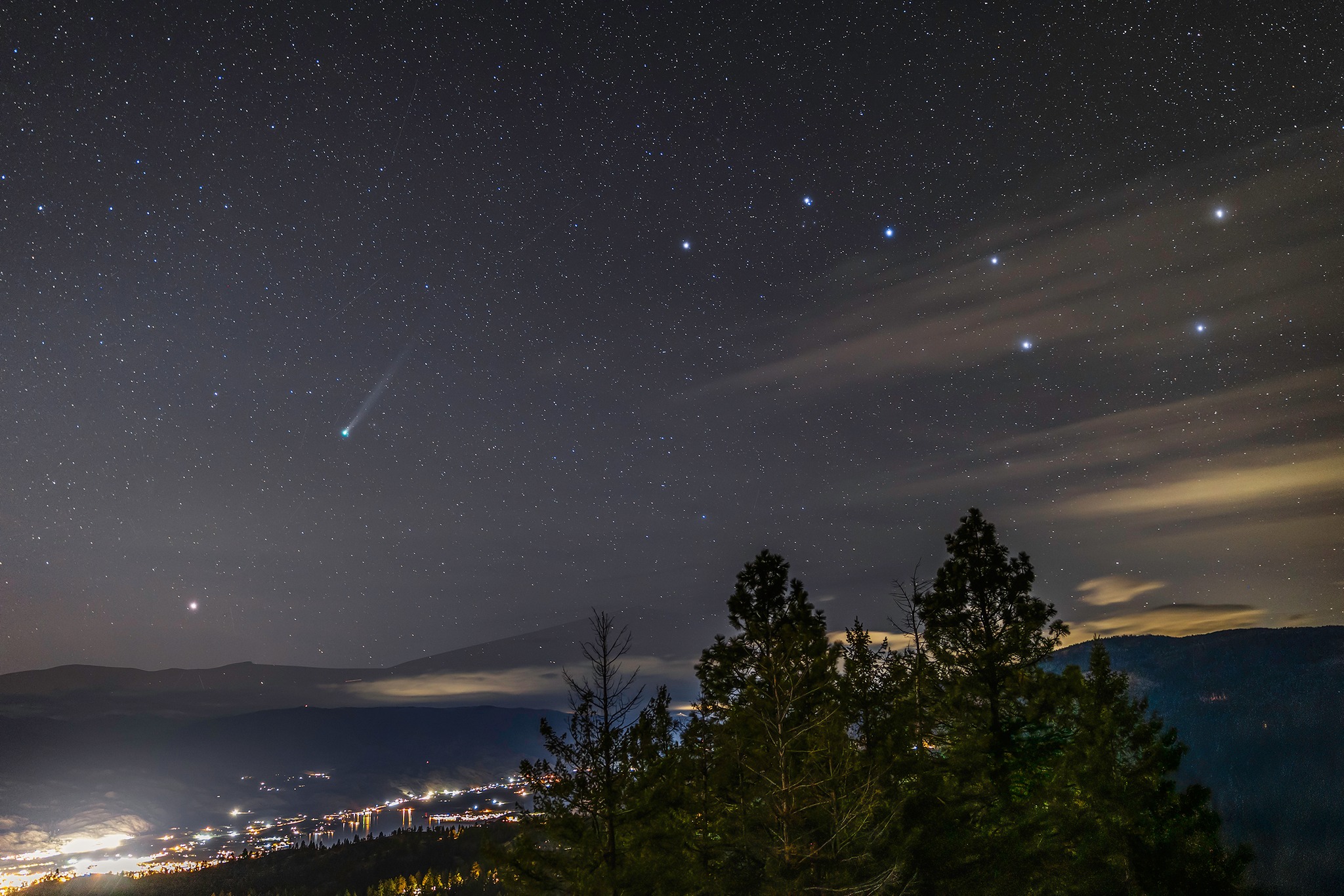 A night sky full of stars, the big dipper and a comet are seen over the distant lights of a town.
