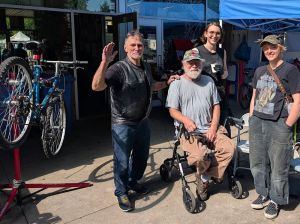 Three people stand around a person in a wheelchair outside a repair shop.