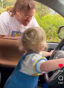 A baby behind the wheel of a car with a man looking in the window.