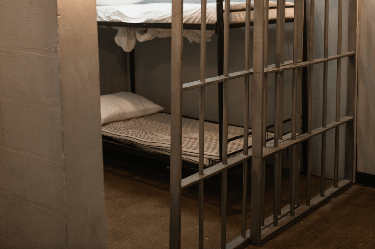 A bed seen through the bars of a prison cell.