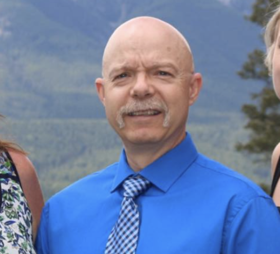 A bald man with a moustache wearing a blue shirt and a tie poses for a photo.