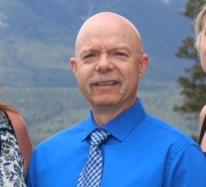 A bald man with a moustache wearing a blue shirt and a tie poses for a photo.