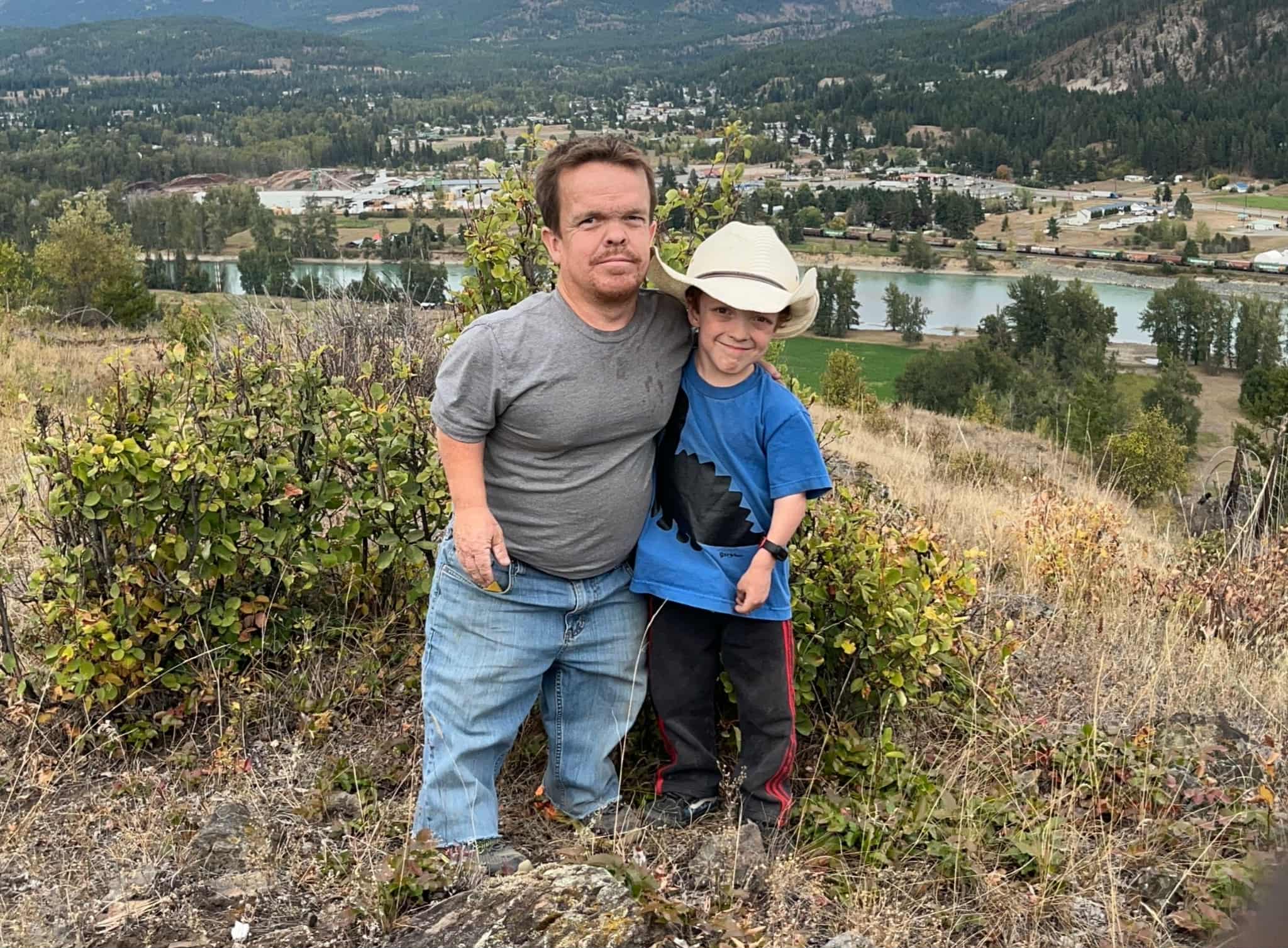 A father and his son pose with their arms around each other on a grassy hill overlooking a river.