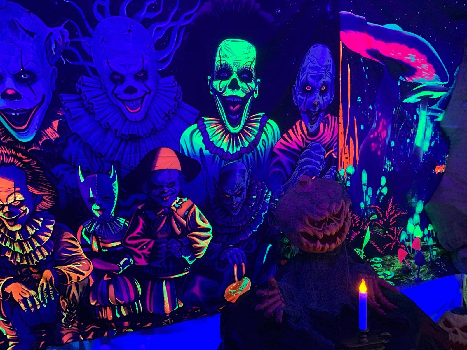 A decorative scary pumpkin head is staged before a lit candle in front of a neon backdrop of clowns.