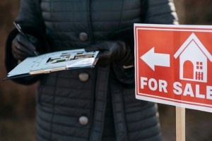 A realtor holds a clipboard by a for sale sign.