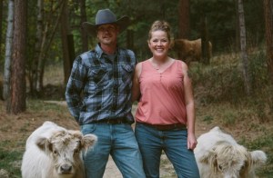 A couple stands on a dirt road in a forest with a miniature cow on either side of them.