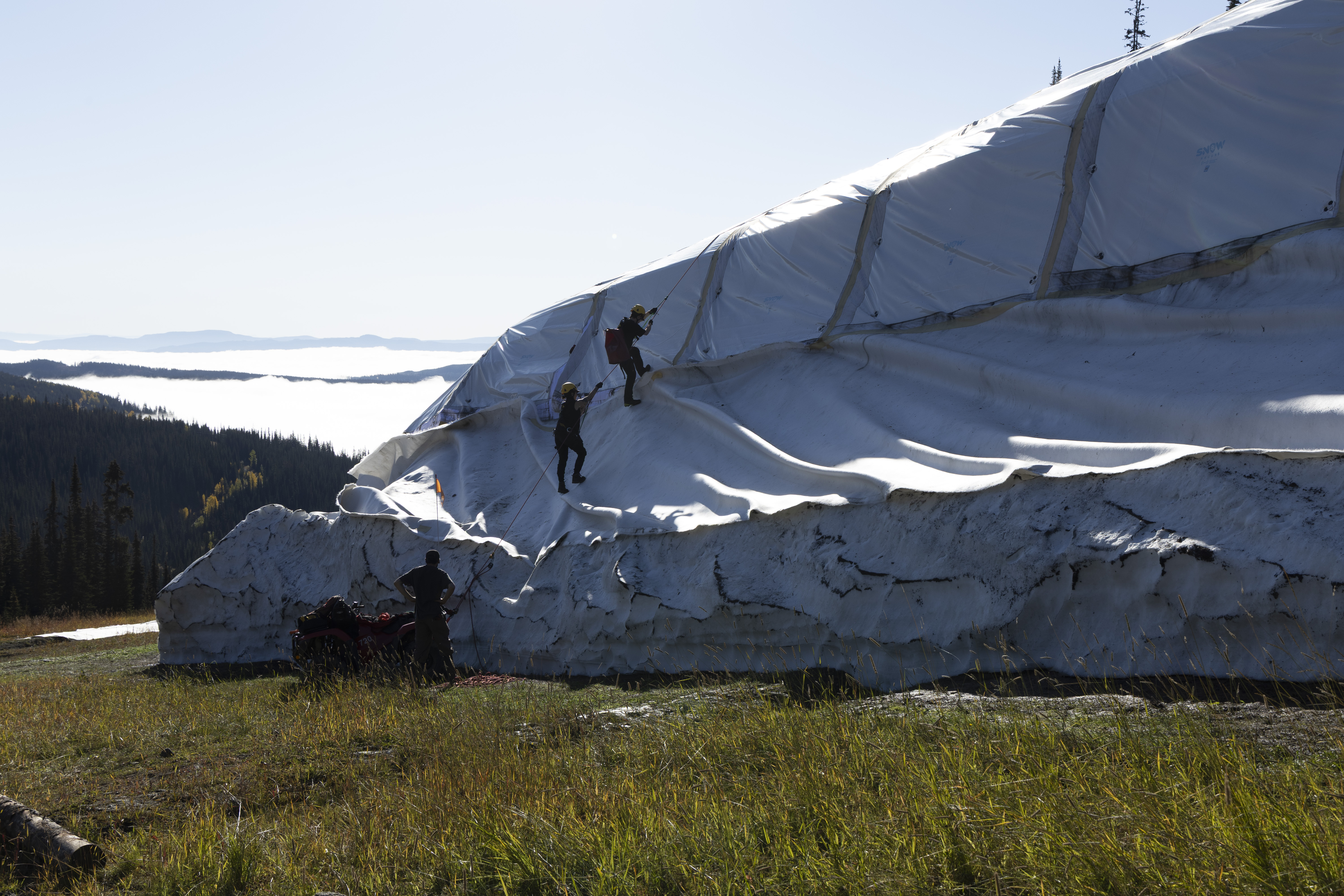 Three people climb a covered pile of snow using a rope.