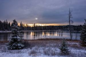 A still mountain lake surrounded by snow dusted trees is seen at sunset.
