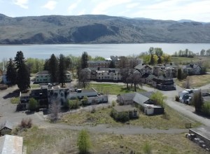 A cluster of buildings off all shapes and sizes sit on flat land among scattered trees with a lake in the background.