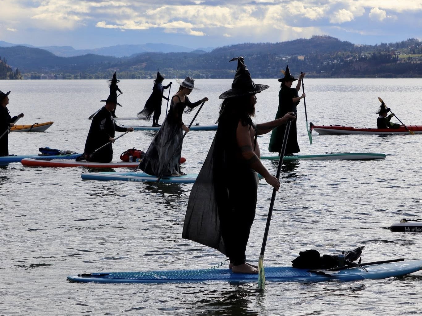 Seven people dresses as witches paddle on boards on a calm lake.